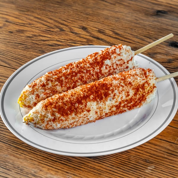 Two elotes, Mexican street corn, covered in white cheese and red chili powder on a white plate.