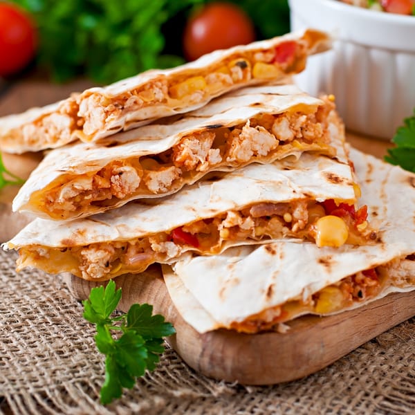 Stacked chicken and corn quesadillas on a wooden board, with tomatoes and parsley in the background.