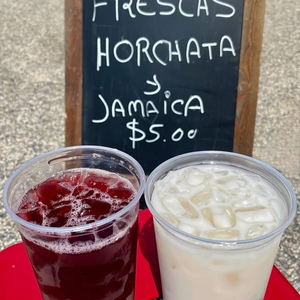 Two drinks, a red Jamaica and a white Horchata, sit in front of a chalkboard menu.