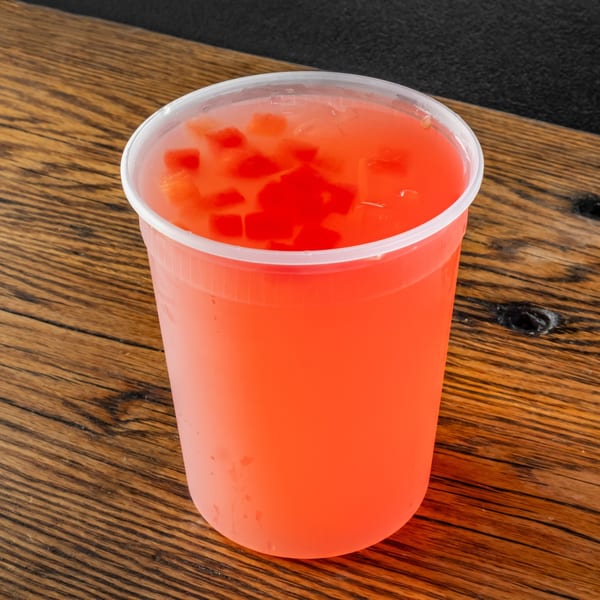 A plastic cup of vibrant red agua fresca with small fruit chunks, on a rustic wooden table.